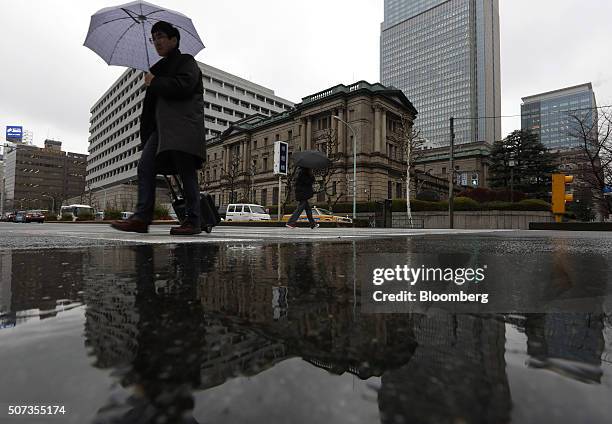 Pedestrians holding umbrellas while walking past the Bank of Japan headquarters are reflected in a puddle in Tokyo, Japan, on Friday, Jan. 29, 2016....