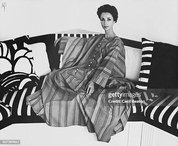 Seated portrait of Gloria Guinness wearing a Mexican striped djellaba with white button down front, seated on a striped couch.