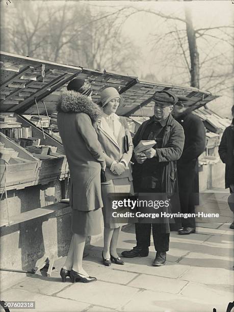 Two models looking at book held by bookseller along the Seine, Paris; model at left wearing velour suit with simple skirt slightly flaring at the...