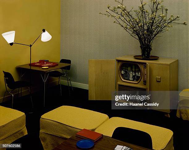 Casual room with a television-phonograph cabinet against the back wall, two yellow ottomans for viewing, a corner table with two black Eames chairs,...