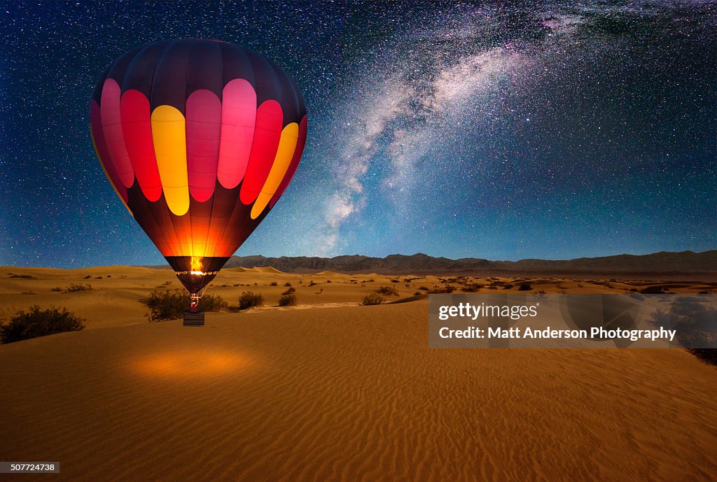 A majestic hot air balloon soars under the stars of the Milky Way, over the desert - Mesquite Dunes of Death Valley National Park. Moonlight provides luminosity showing the patterns and shapes of the desert landscape.