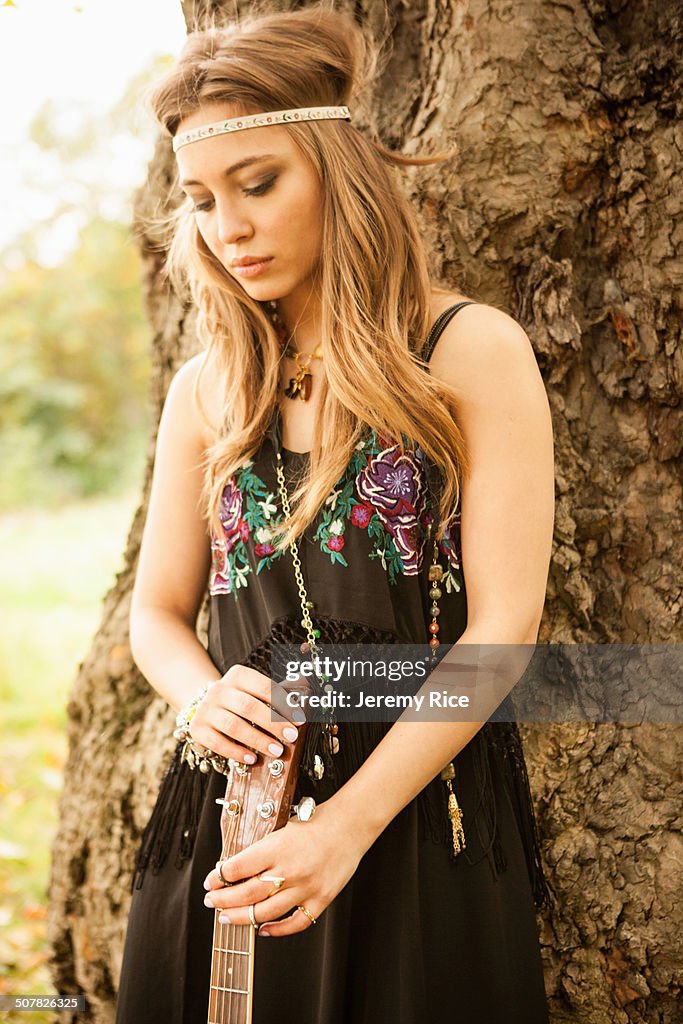 Hippy woman with guitar looking sad standing by tree