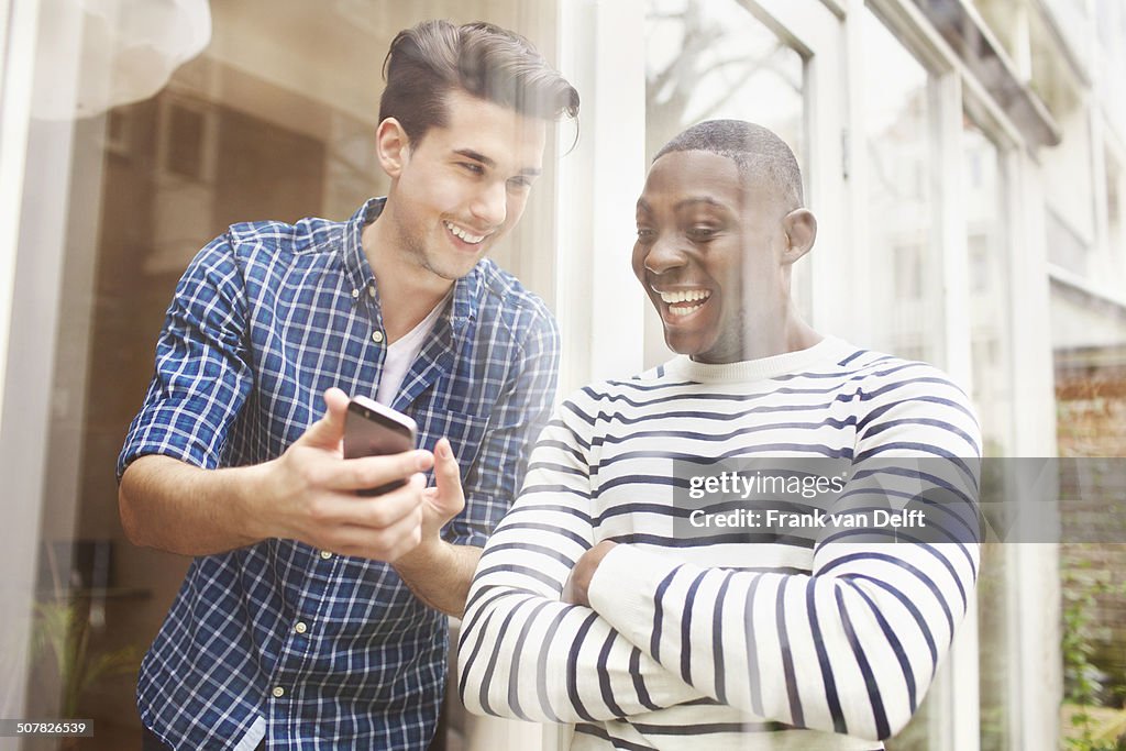 Two young male friends looking at smartphone behind patio glass