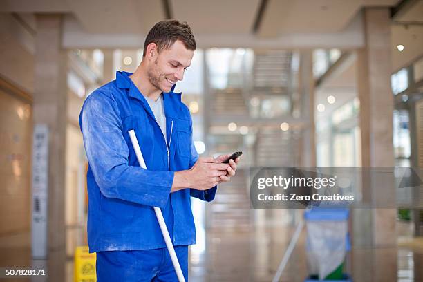 male cleaner texting on smartphone in office atrium - schoonmaker man stockfoto's en -beelden