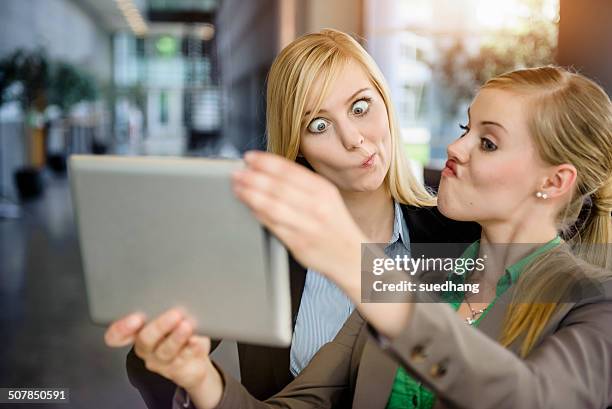 two young businesswomen pulling faces and taking selfie on digital tablet in office - münchen business stock pictures, royalty-free photos & images