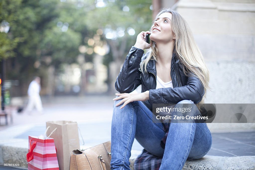 Young woman shopper sitting on city steps chatting on smartphone