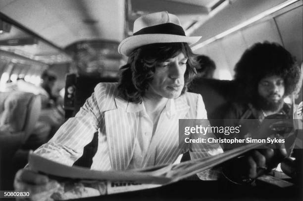Mick Jagger keeps abreast of current affairs while travelling between concerts in the Rolling Stones' private jet during their 1975 Tour of the...