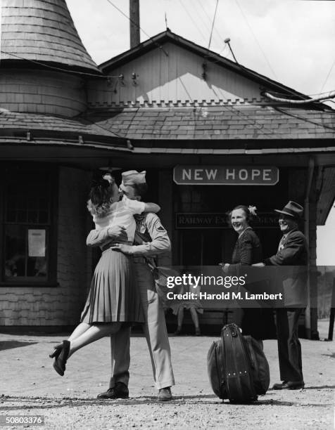 At a train station, a soldier lifts a woman off her feet as he kisses her, while a couple watches, 1940s.