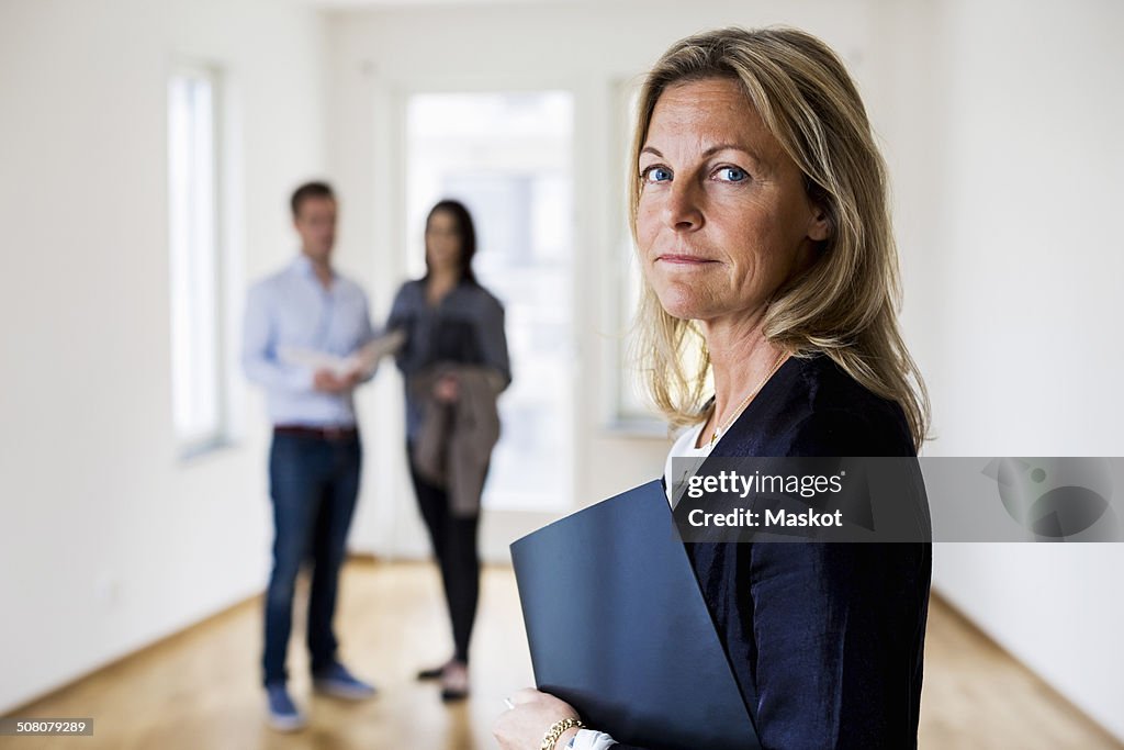 Portrait of confident female real estate agent with couple standing in background at home