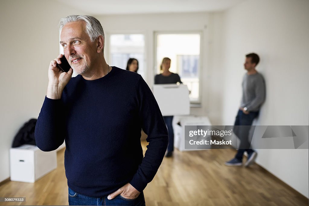Mature man using mobile phone while family with moving boxes in background at home