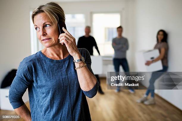 mature woman using mobile phone while family carrying moving boxes in background at home - mother in law stock pictures, royalty-free photos & images