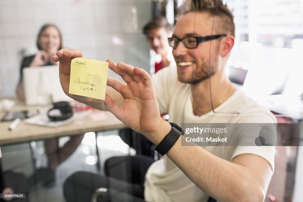 Young businessman sticking adhesive note on transparent glass with colleagues at background in new office