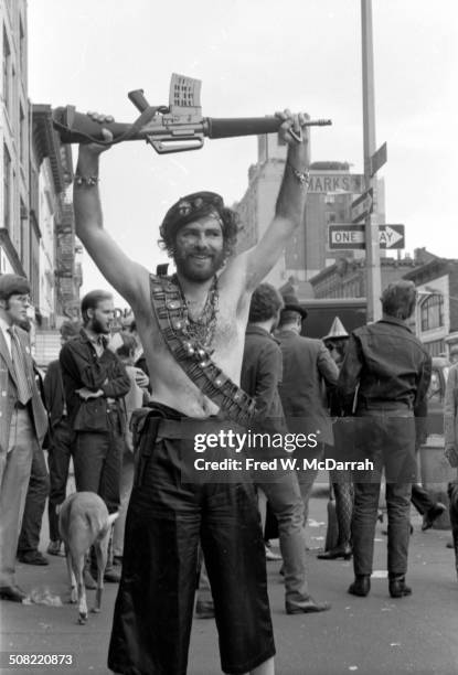 American social and political activist Jerry Rubin stands shirtless as a he holds an M16 assault rifle above his head at the intersection of St Marks...