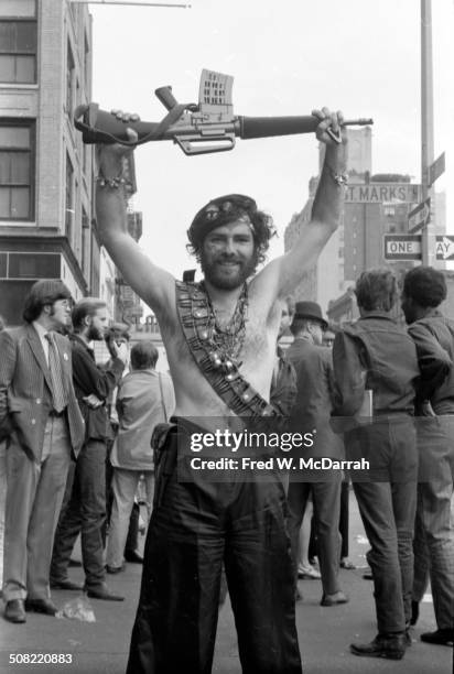 American social and political activist Jerry Rubin stands shirtless as a he holds an M16 assault rifle above his head at the intersection of St Marks...