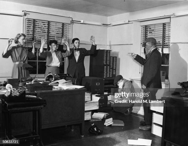 Masked and armed robber holds the staff of an office at gunpoint while his accomplice searches through files, 1940s.