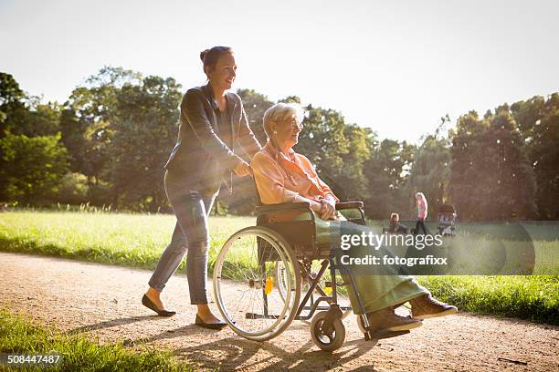 young woman pushing senior lady in wheelchair through a park - övervakning bildbanksfoton och bilder