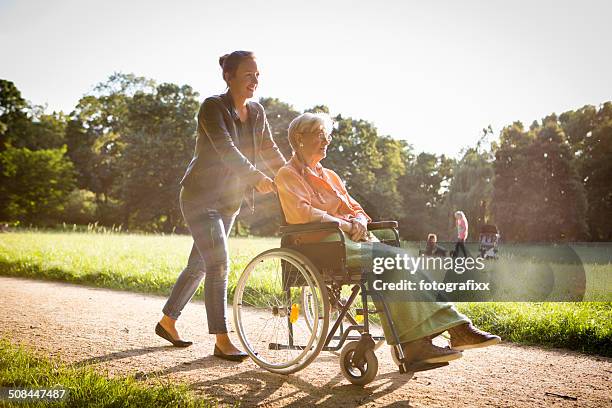 young woman pushing senior lady in wheelchair through a park - 遊園地 個照片及圖片檔