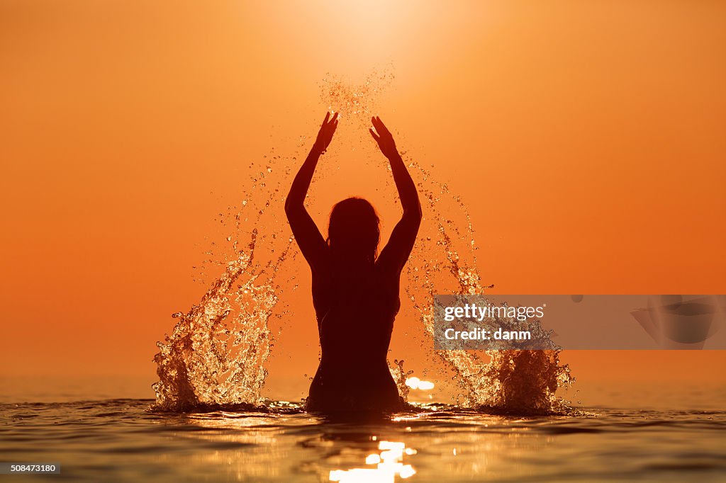 Beauty Model Girl Splashing Water with her hands. Teen girl Swimming and splashing on summer beach over sunset. Beautiful Woman in Water