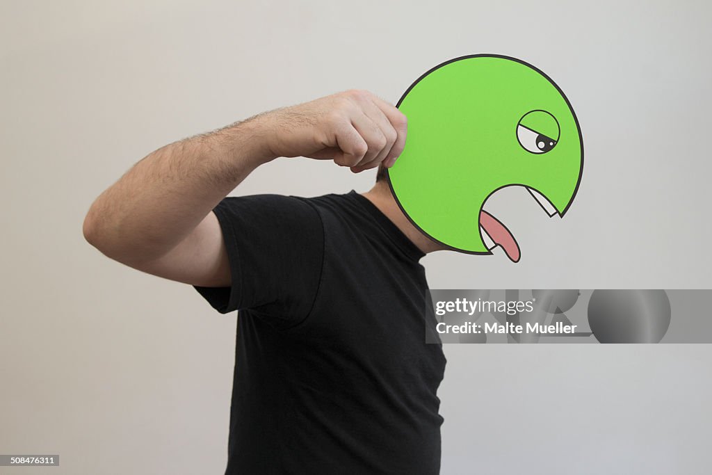 Side view of man holding a green vomit emoticon face in front of his face