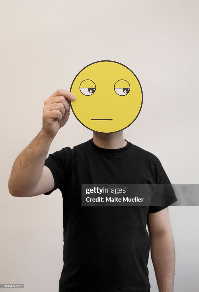 Man holding a bored emoticon face in front of his face