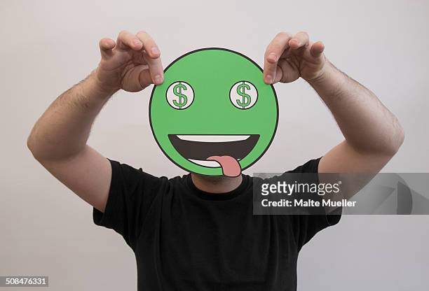 man holding a green dollar sign emoticon face in front of his face - stick out tongue emoji stock-fotos und bilder