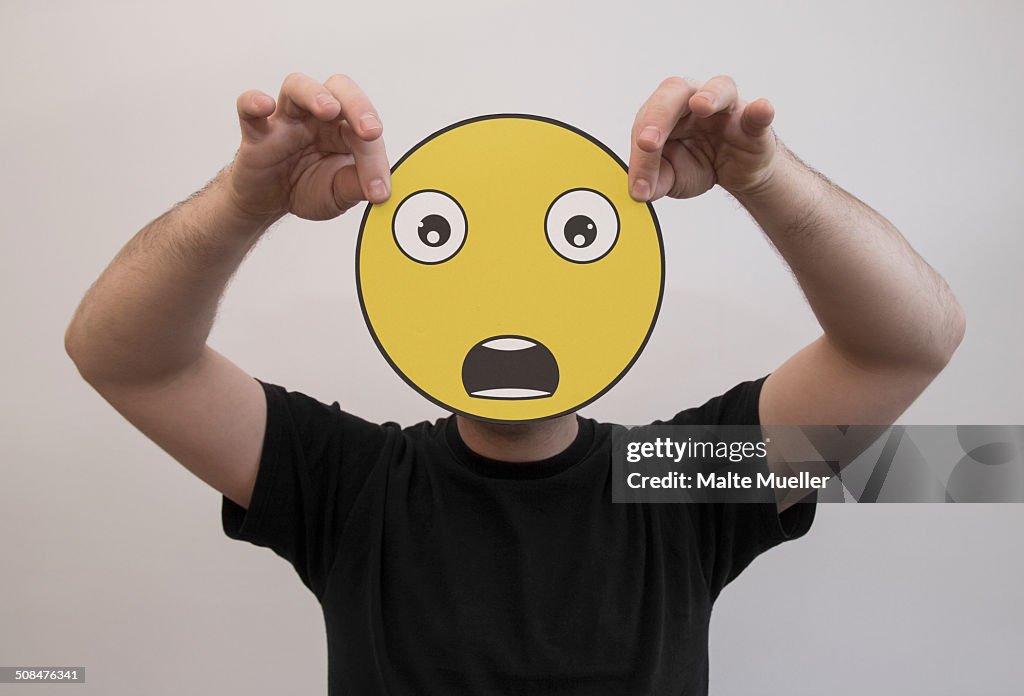 Man holding a shocked emoticon face in front of his face