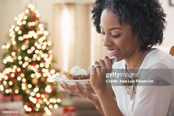 mixed race woman eating dessert by christmas tree - christmas dessert stock pictures, royalty-free photos & images