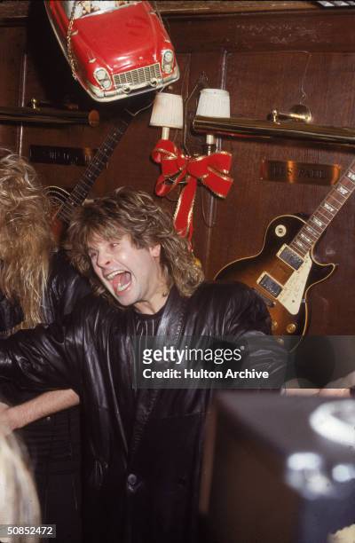 British heavy metal singer Ozzy Osbourne screams inside a bar, with an electric guitar mounted on the wall behind him, 1980s.
