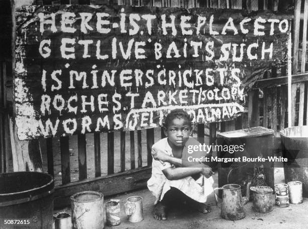 African-American child crouching between various cans in front of sign while selling live bait.