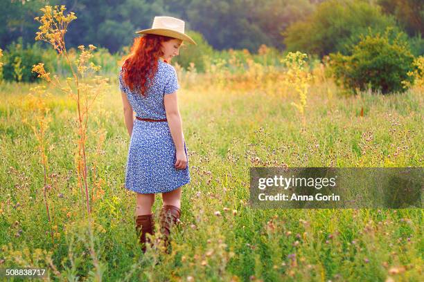 redhead in cowboy hat and dress in sunlit field - cowgirl hairstyles stock-fotos und bilder