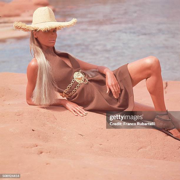 Verushka reclining in the red Arizona desert in a knit dress by Lotte with a gold chain link Palacio medallion belt, worn with a wide brimmed Adolfo...