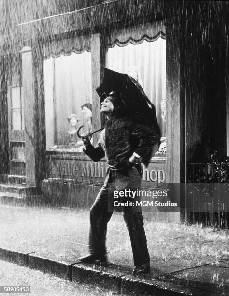 American actor Gene Kelly stands on a sidewalk smiling and holding an umbrella during a downpour in a still from the film 'Singin' in the Rain',...