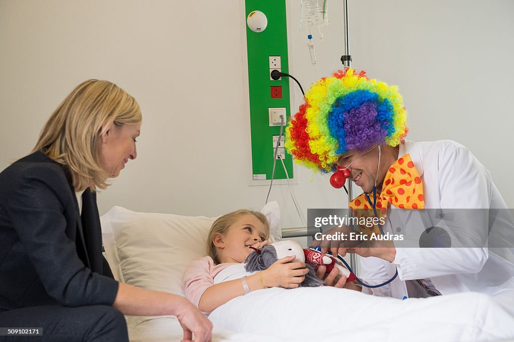 Male doctor wearing clown costume making girl patient smile in hospital bed