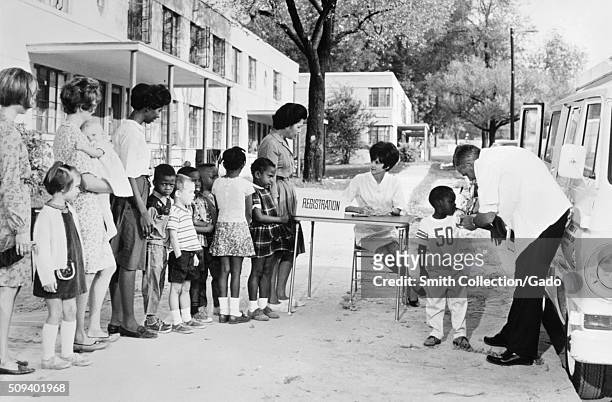This 1976 photograph depicts a multiracial group of women and children in a housing project lining up at a mobile clinic waiting for, and receiving...
