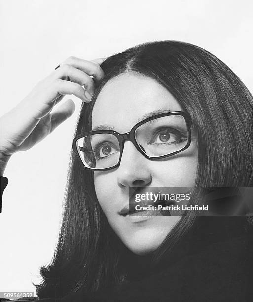 Portrait of Greek singer Nana Mouskouri wearing dark-framed rectangular glasses, one hand pulling hair back from her face.