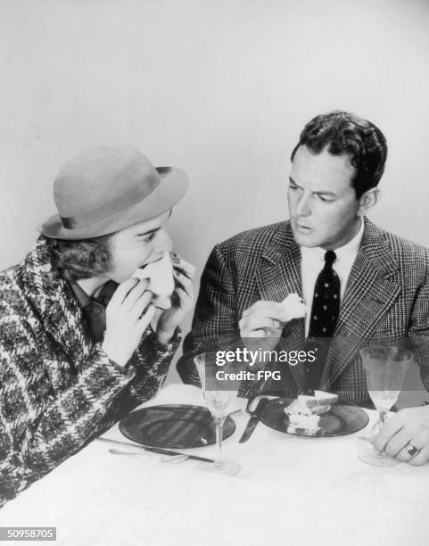 Man expresses disapproval at his friend's table manners as she sinks her teeth greedily into a sandwich, circa 1940.