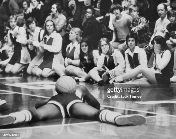 Spectators and cheerleaders near courtside react in distress as an unidentified basketball player, with a ball on his back, lies prone on the court,...