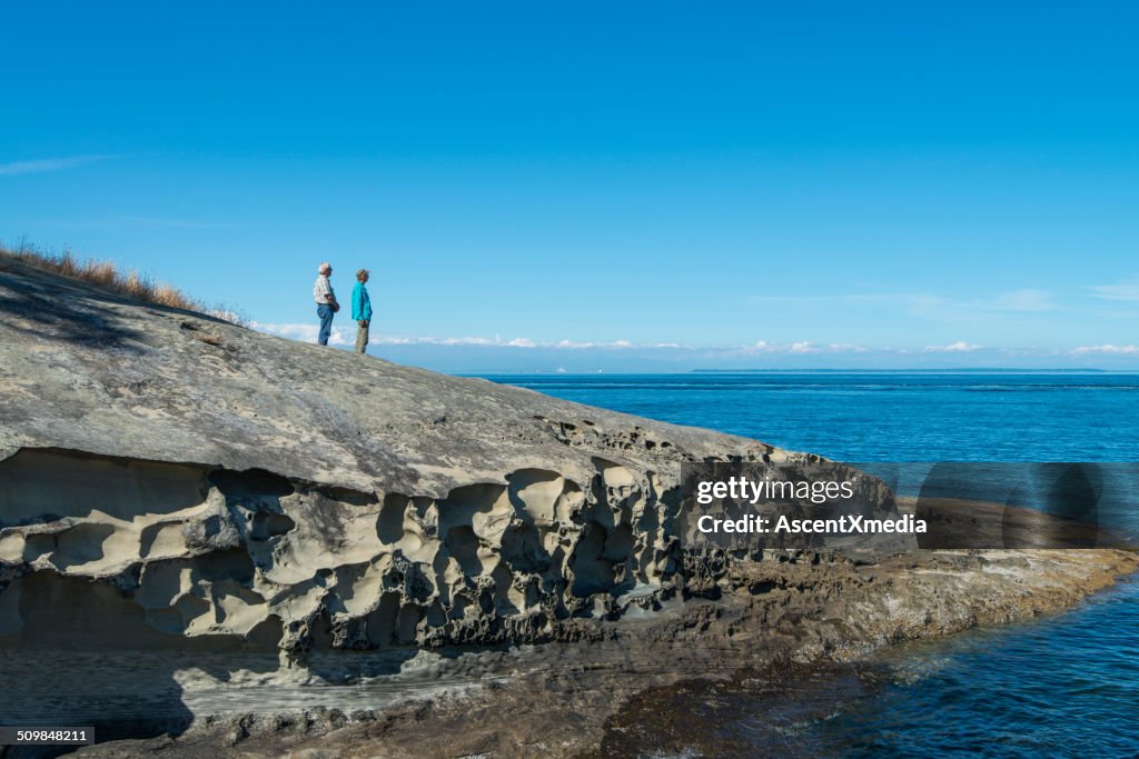 Senior couple stand on coastal rocks, look to sea
