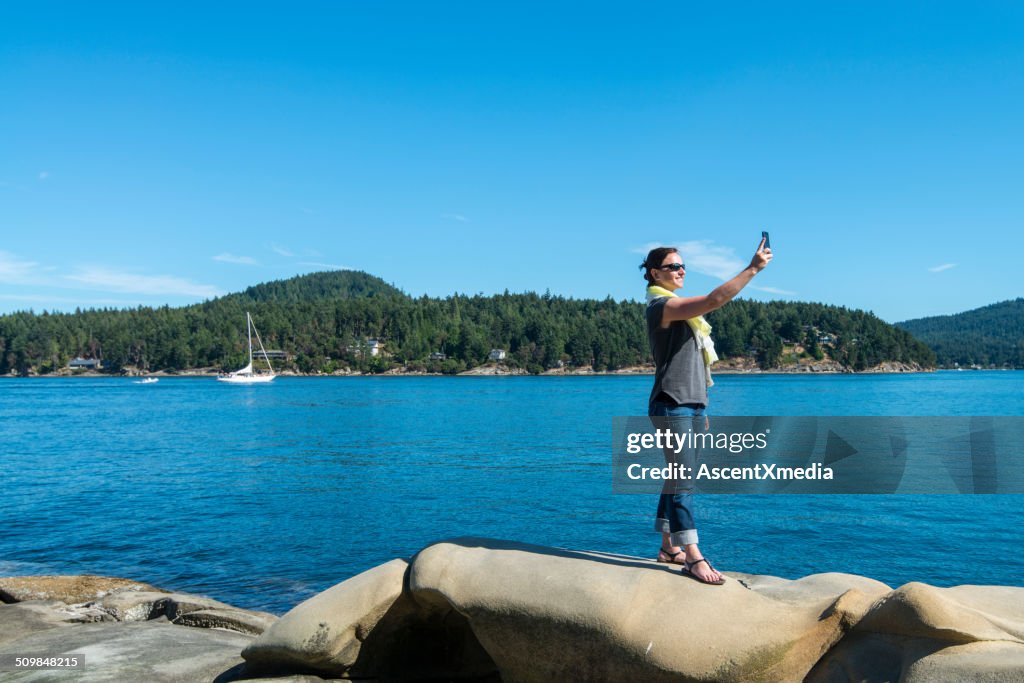 Woman stands on rocky shoreline, takes selfie