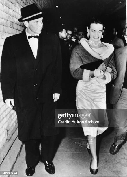 American pop singer and actor Frank Sinatra in an overcoat and hat walks next to American heiress and designer Gloria Vanderbilt as she wears a...