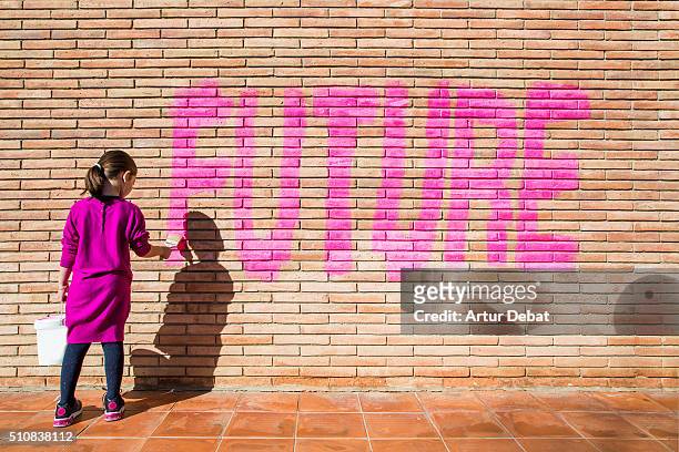 little girl painting with pink colors the future word in a brick wall, a protest action claiming for future to the new generations. - learning objectives fotografías e imágenes de stock