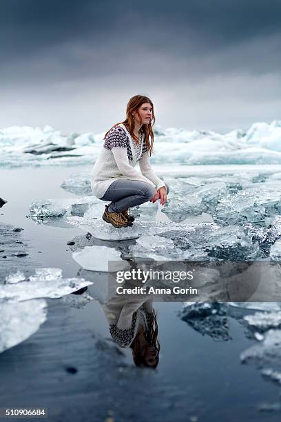 young woman crouches on iceberg at jokulsarlon glacial lagoon, reflected in water - jokulsarlon lagoon fotografías e imágenes de stock