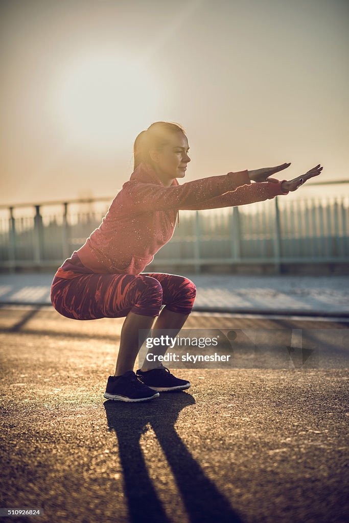 Athletic woman doing squats on a road at sunset.