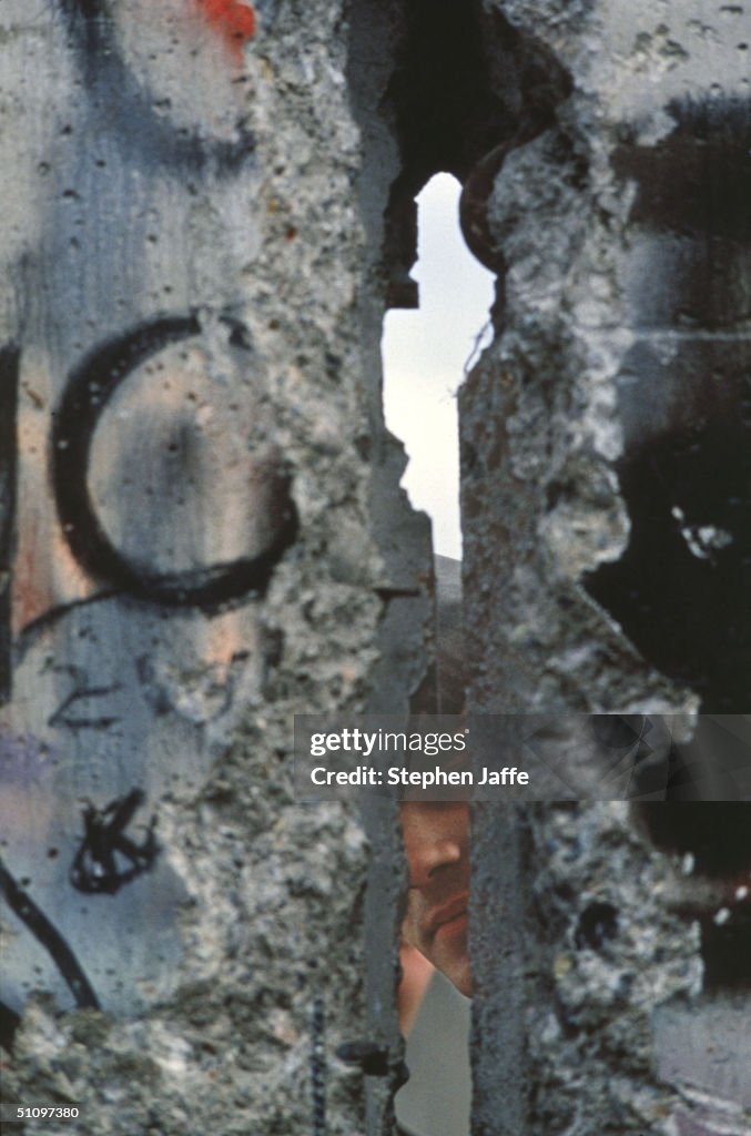 An East German Border Guard Peers Through A Broken Piece Of The Berlin Wall During The C...