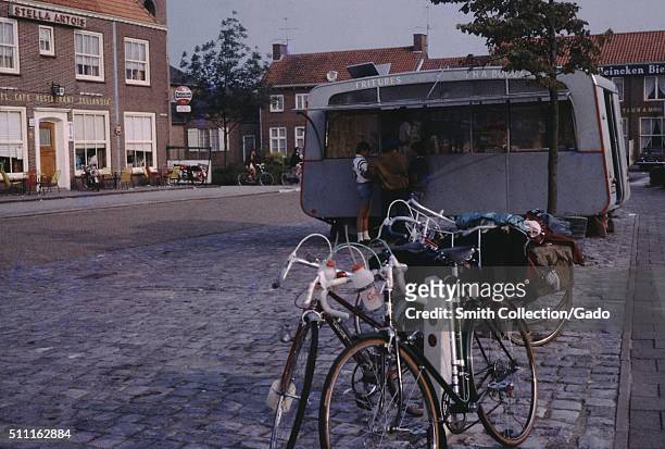 Bicycles parked outside a french fry stand in a public square, with a cafe and a Stella Artois beer advertisement, Holland, 1960. .