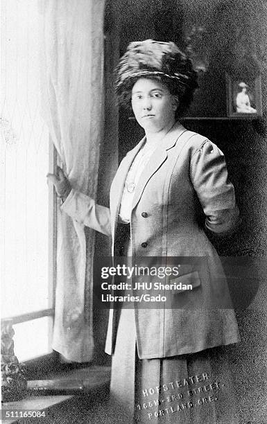 Portrait of an African American woman standing next to a window, wearing a hat, glasses and a two-piece jacket and skirt, with a framed photograph of...