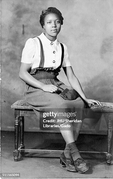 Portrait of an African American woman sitting on a bench, wearing a white button down shirt and a high-waisted skirt, holding a purse, 1915. .