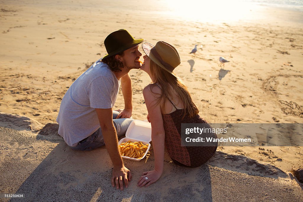 Hipster couple feeding eachother chips on Australian beach at sunset