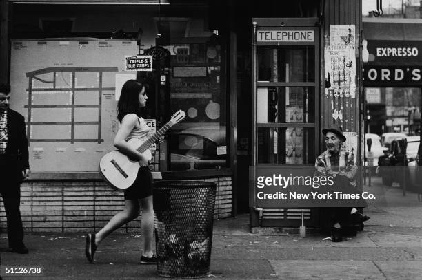 Young woman walks down a sidewalk in Greenwich Village, carrying an acoustic guitar, New York City, April 25, 1961.