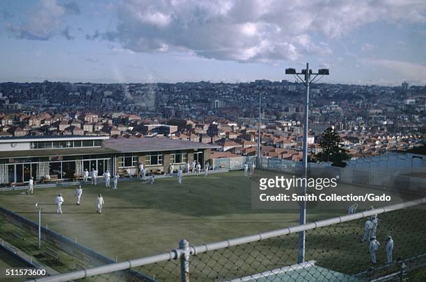 Photograph taken from an elevated viewpoint of a cricket match being played, a clubhouse can be seen in the background and benches for viewers can be...
